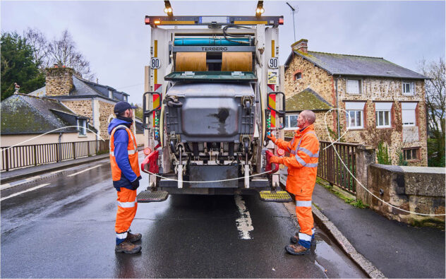 2 rippeurs sont à l'arrière d'une benne à ordures. Un bac est en train de se vider dans celle-ci.