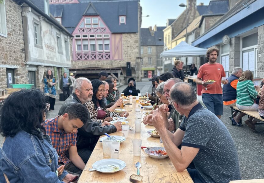 Plusieurs personnes de tout âge sont attablées et partagent un repas dans une rue de Lamballe-Armor. C'est un moment festif et de partage.
