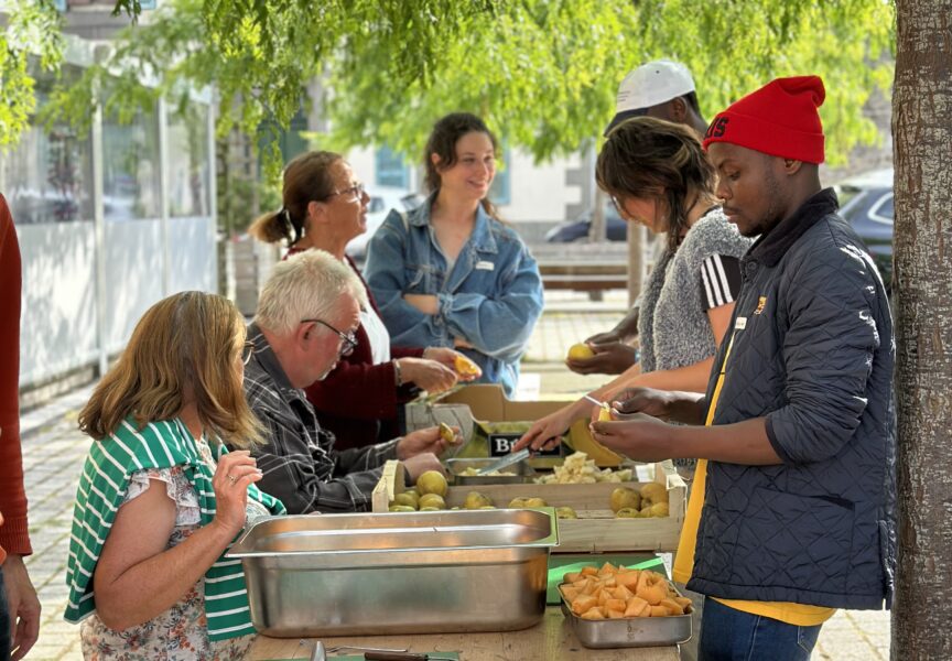 Un groupe de personnes, des jeunes et des séniors, épluchent et découpent des légumes autour d'une table, situé sous un arbre. Des sourires sont visibles sur les visages.